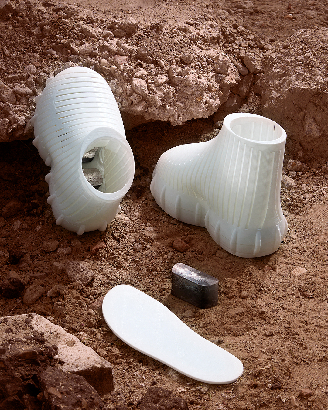 Futuristic, white running shoes standing on red, rocky soil. Palm sized blocks of metal can be seen on the inside of the left shoe. A block of metal and the white inner sole of the left shoe are laying on the ground.